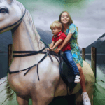 Children riding a horse on a lakeside dock surrounded by misty mountains